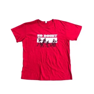 Puede incluir: Camiseta roja con un gráfico en blanco y negro de una banda y el texto "NO DOUBT" en un rectángulo blanco. El gráfico muestra siluetas de músicos tocando instrumentos. La camiseta está hecha de un material suave.