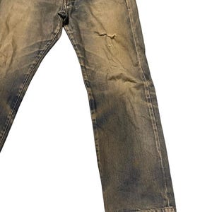 May include: Faded blue denim jeans with a distressed look. The jeans have a button fly and a small tear on the right leg. The denim has a worn appearance with lighter areas.