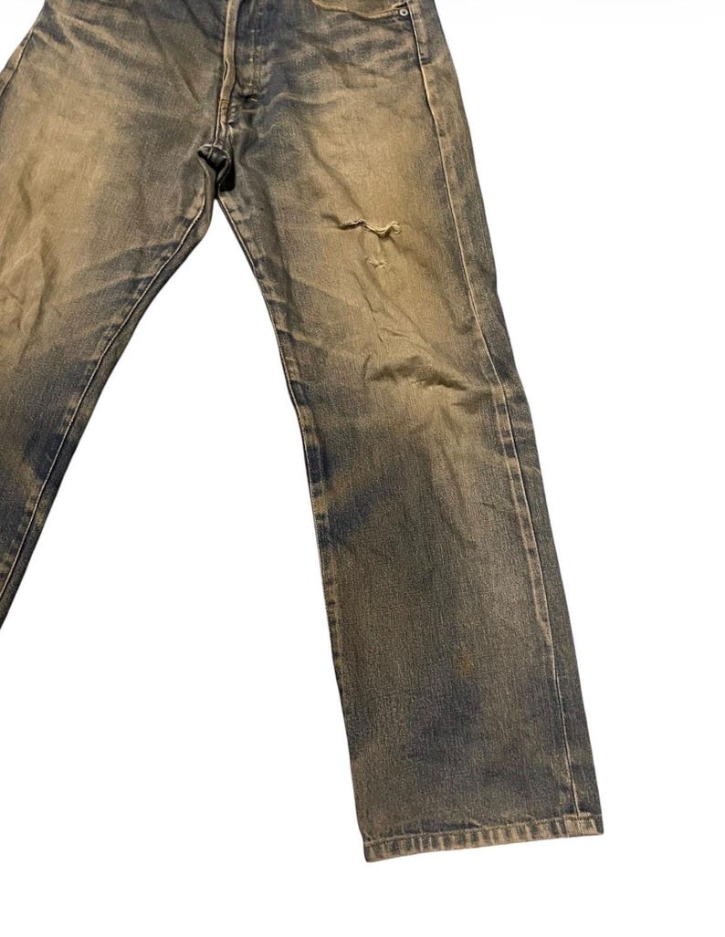 May include: Faded blue denim jeans with a distressed look. The jeans have a button fly and a small tear on the right leg. The denim has a worn appearance with lighter areas.