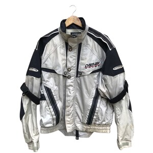 K&ouml;nnte beinhalten: Eine silberne und schwarze Jacke mit Rei&szlig;verschluss und einem CYBER-TEX-Logo auf der Vorderseite. Die Jacke hat ein schwarz-wei&szlig;es Streifenmuster an den &Auml;rmeln und einen schwarzen Kragen.
