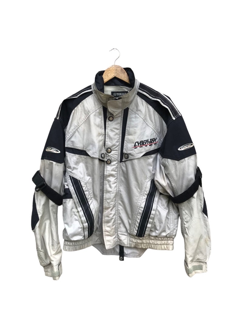 K&ouml;nnte beinhalten: Eine silberne und schwarze Jacke mit Rei&szlig;verschluss und einem CYBER-TEX-Logo auf der Vorderseite. Die Jacke hat ein schwarz-wei&szlig;es Streifenmuster an den &Auml;rmeln und einen schwarzen Kragen.