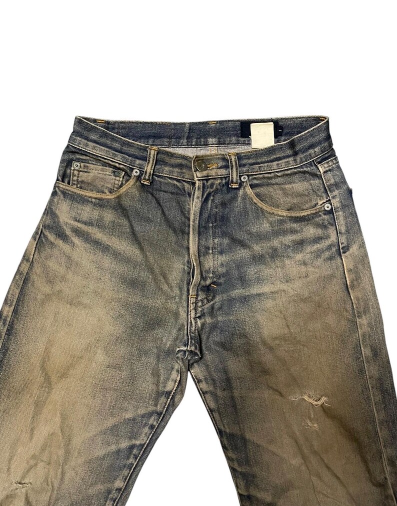 May include: Faded blue denim jeans with a worn appearance. The jeans have a classic five-pocket design, a button closure, and a distressed look with a small tear on the leg. The denim has a vintage wash.