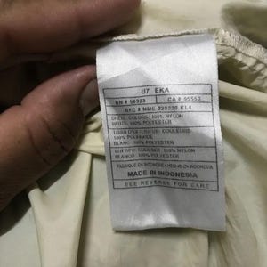 May include: Close-up of a tag with text. The tag is white with black text and is attached to a cream-colored fabric. The tag includes information about the shell colors, materials, and manufacturing location.