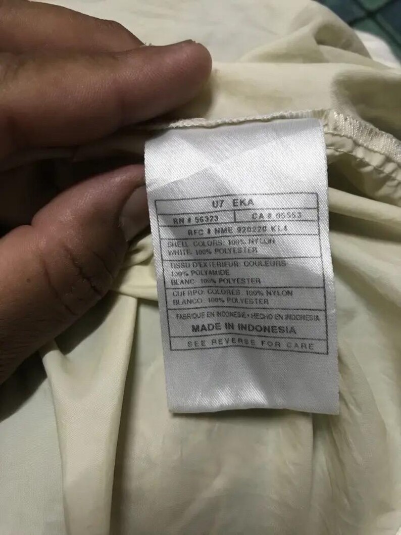 May include: Close-up of a tag with text. The tag is white with black text and is attached to a cream-colored fabric. The tag includes information about the shell colors, materials, and manufacturing location.