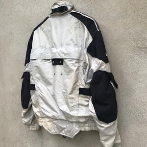 K&ouml;nnte beinhalten: Eine wei&szlig;e und schwarze Jacke mit Rei&szlig;verschluss und mehreren Taschen. Die Jacke hat einen reflektierenden Streifen am &Auml;rmel.