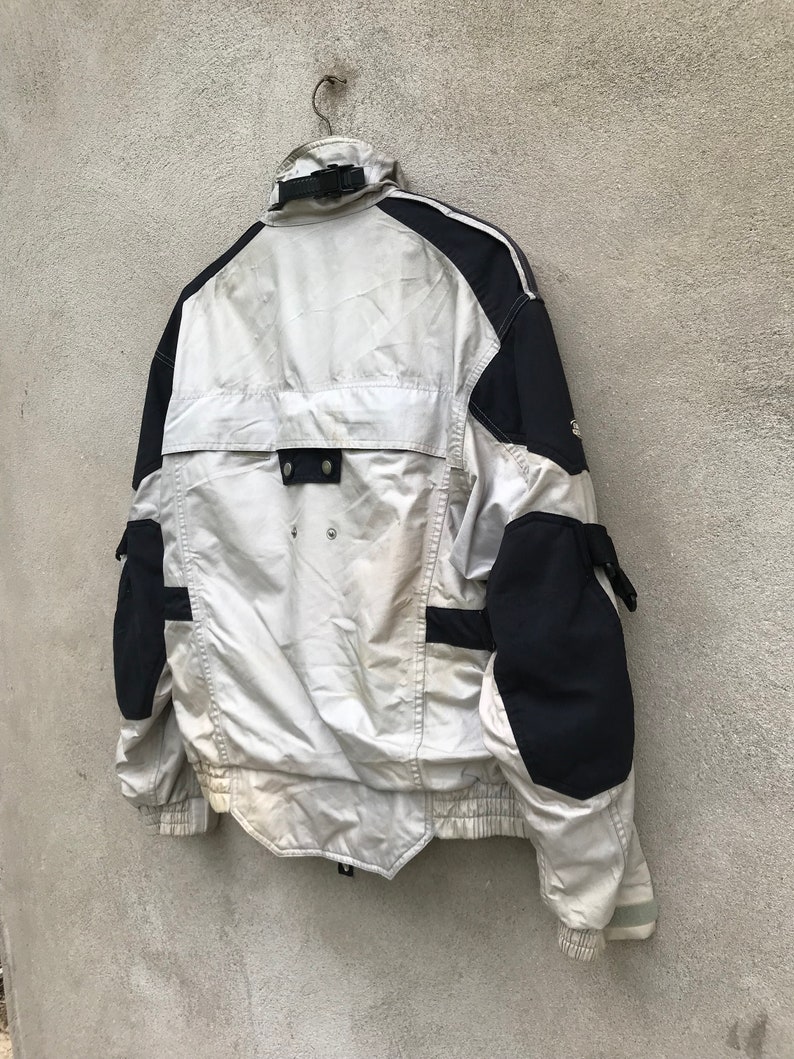 K&ouml;nnte beinhalten: Eine wei&szlig;e und schwarze Jacke mit Rei&szlig;verschluss und mehreren Taschen. Die Jacke hat einen reflektierenden Streifen am &Auml;rmel.
