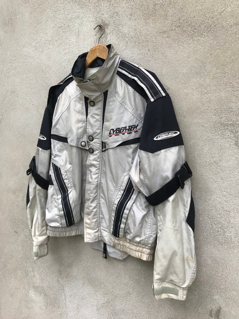 K&ouml;nnte beinhalten: Eine silberne und schwarze Jacke mit dem Aufdruck "CYBER-TECH" auf der Vorderseite. Die Jacke hat schwarze Streifen und einen Rei&szlig;verschluss.
