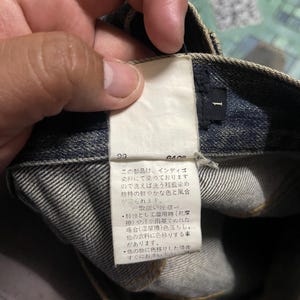 May include: Close-up of a pair of blue denim jeans with a white tag attached. The tag has Japanese text and the number "1" printed on it. The jeans are folded, showing the inside of the waistband.
