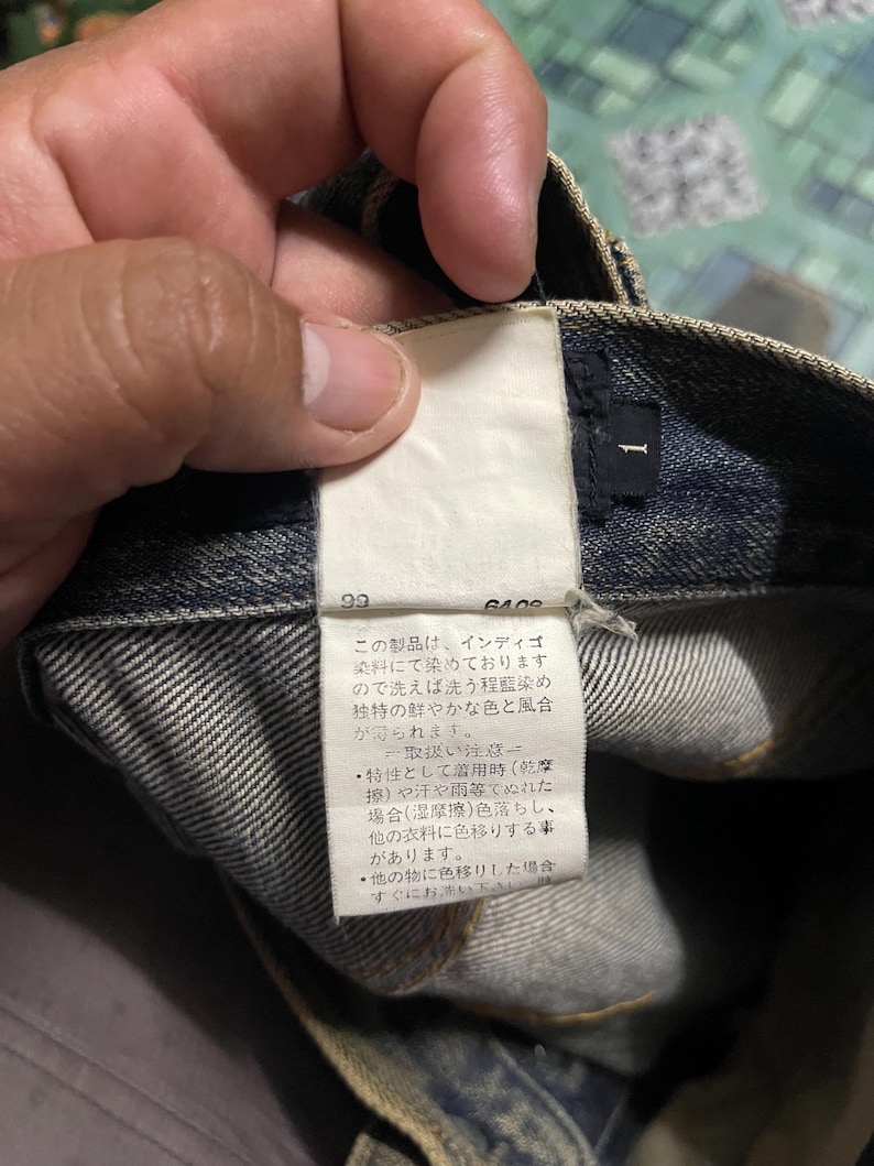 May include: Close-up of a pair of blue denim jeans with a white tag attached. The tag has Japanese text and the number "1" printed on it. The jeans are folded, showing the inside of the waistband.