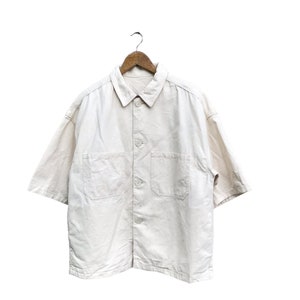 Può includere: Una camicia bianca a maniche corte con bottoni e due tasche sul petto, appesa a un appendiabiti in legno.