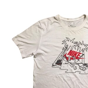 Op de afbeelding: Wit T-shirt met een afbeelding van een paar Nike sneakers in een hangmat tussen twee palmbomen. De tekst "NIKE" staat in rood en zwart op de sneakers. De tekst "THE NIKE TEE" staat op het label.