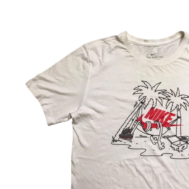 Op de afbeelding: Wit T-shirt met een afbeelding van een paar Nike sneakers in een hangmat tussen twee palmbomen. De tekst "NIKE" staat in rood en zwart op de sneakers. De tekst "THE NIKE TEE" staat op het label.