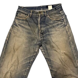 May include: Faded blue denim jeans with a distressed appearance. The jeans have a button fly, classic five-pocket design, and a worn-in look with a small tear on the right leg. The denim has a vintage wash.