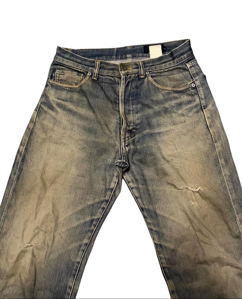 May include: Faded blue denim jeans with a distressed appearance. The jeans have a button fly, classic five-pocket design, and a worn-in look with a small tear on the right leg. The denim has a vintage wash.