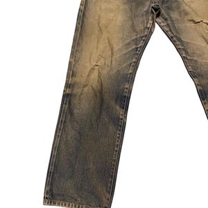 May include: Faded blue denim jeans with a distressed appearance. The jeans have a button fly and show signs of wear, including a small tear on the right leg. The denim has a vintage, worn look.