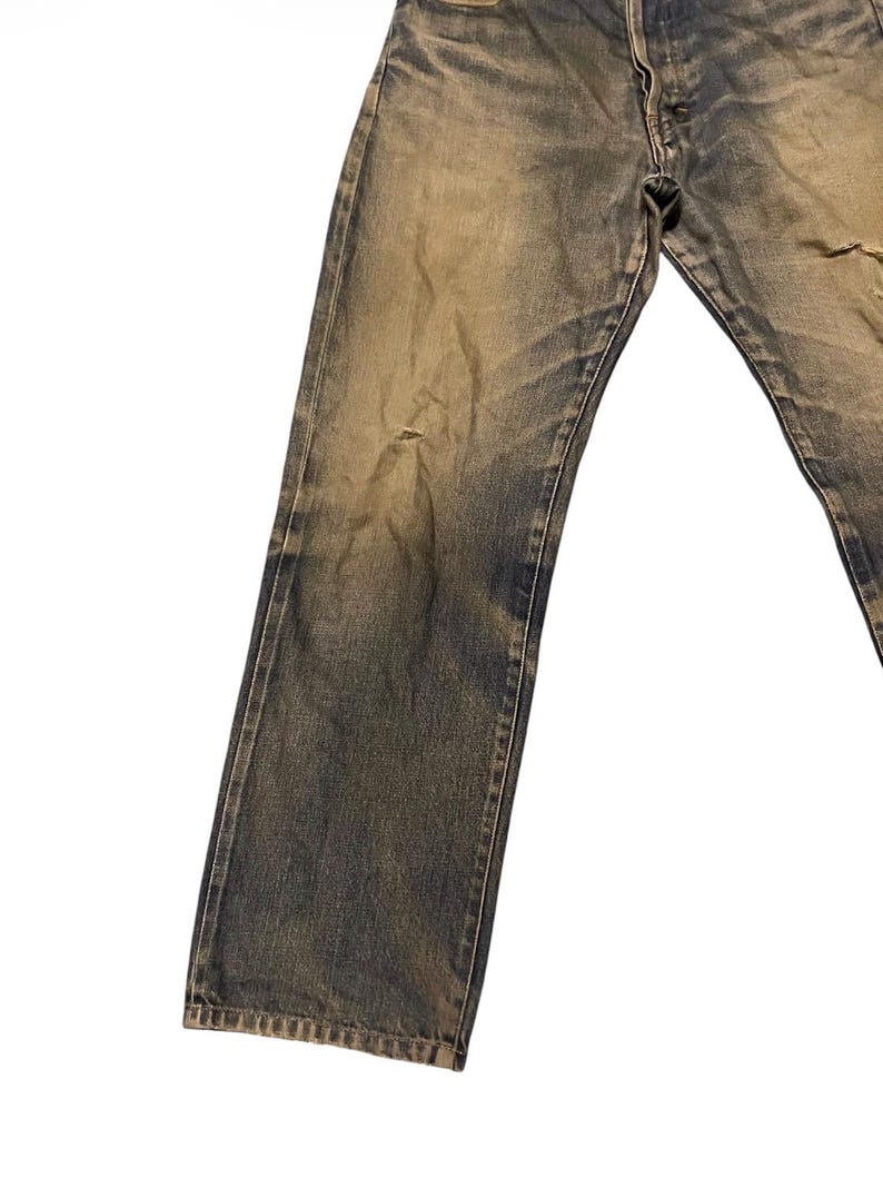 May include: Faded blue denim jeans with a distressed appearance. The jeans have a button fly and show signs of wear, including a small tear on the right leg. The denim has a vintage, worn look.