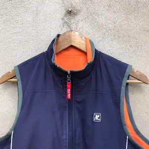 May include: A navy blue vest with a zipper closure and orange lining. The vest has a white logo on the front that says "AC".