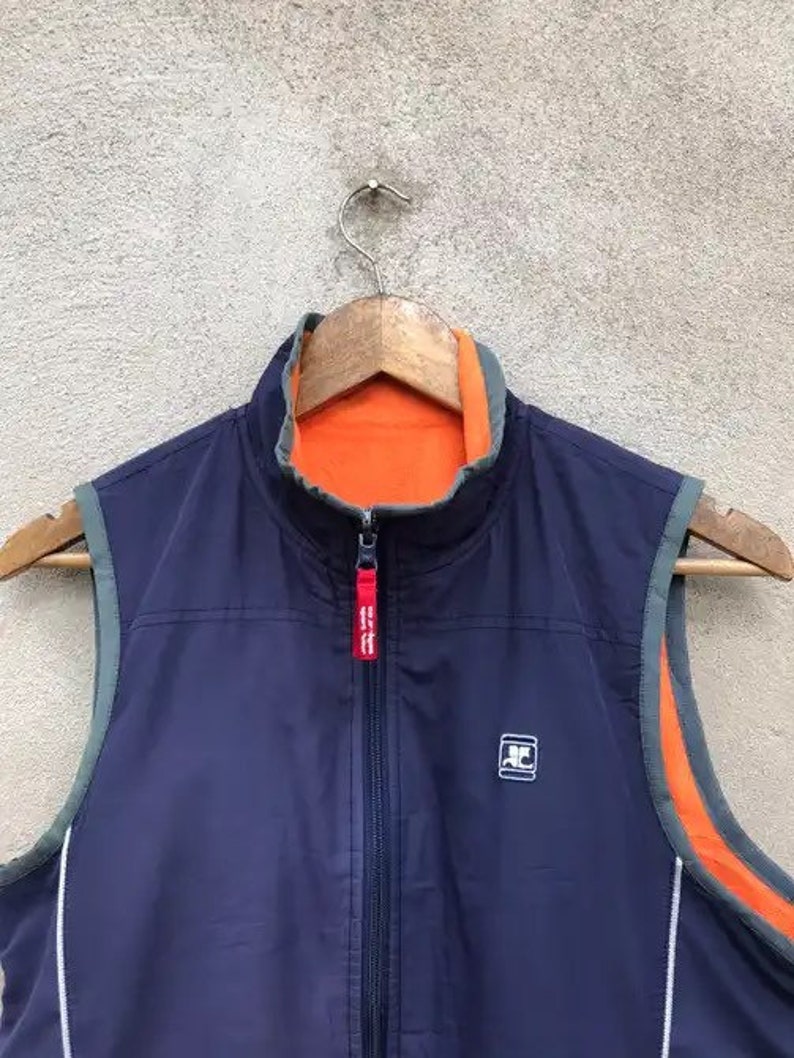 May include: A navy blue vest with a zipper closure and orange lining. The vest has a white logo on the front that says "AC".