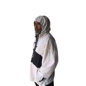 May include: A hooded anorak jacket in white, black, and cream. The jacket features a color-block design with a black panel across the chest and a white hood. The jacket has a zipper closure and pockets. The model is wearing black pants.
