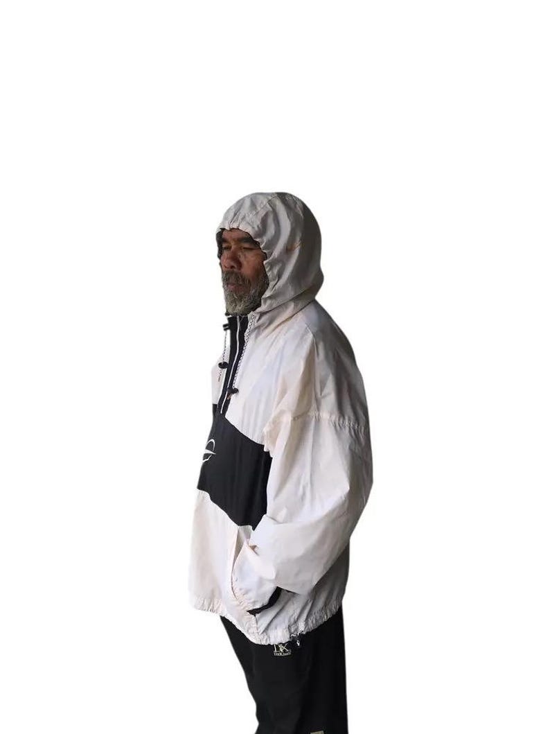 May include: A hooded anorak jacket in white, black, and cream. The jacket features a color-block design with a black panel across the chest and a white hood. The jacket has a zipper closure and pockets. The model is wearing black pants.