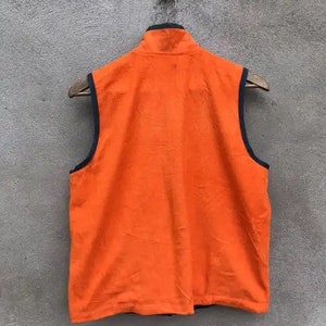 May include: Orange fleece vest with a black zipper and black trim around the armholes. The vest is hanging on a wooden hanger against a gray wall.