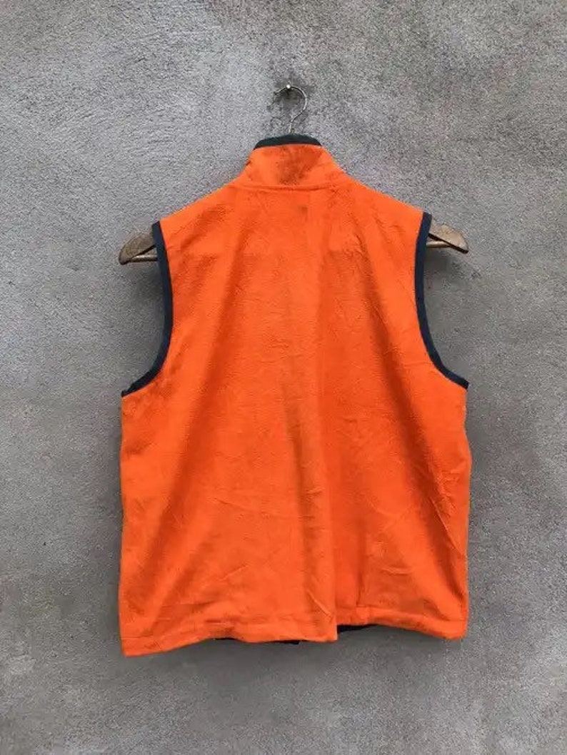 May include: Orange fleece vest with a black zipper and black trim around the armholes. The vest is hanging on a wooden hanger against a gray wall.