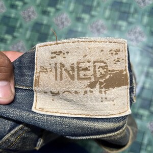 May include: Close-up of a denim jeans label with the word "PINED" printed on it. The label is rectangular, beige, and appears worn. The jeans are a dark blue color.