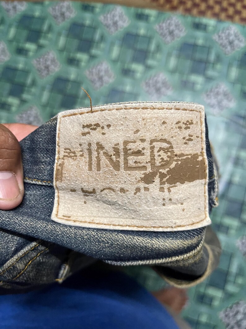 May include: Close-up of a denim jeans label with the word "PINED" printed on it. The label is rectangular, beige, and appears worn. The jeans are a dark blue color.