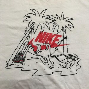 Op de afbeelding: Wit T-shirt met een zwart-witte illustratie van een Nike swoosh in een hangmat tussen twee palmbomen. De Nike swoosh is rood en de tekst "NIKE" is in rode letters. Er is een zwart-witte illustratie van een paar sneakers en een doos met het woord "NIKE" erop.
