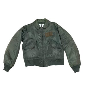 Può includere: Giacca bomber verde oliva con zip, due tasche frontali e una piccola toppa sul petto. La giacca ha polsini a coste e colletto. La giacca è in stile militare.