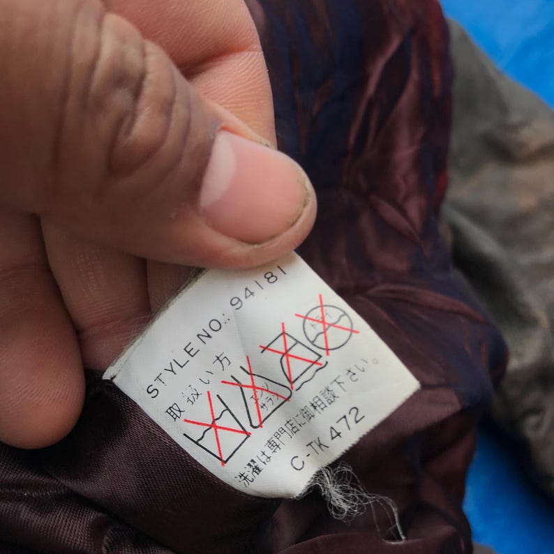 以下が含まれることがあります： 「STYLE NO.: 94181」と「C-TK 472」という白い文字が書かれた、濃い赤紫色の衣服タグのクローズアップ。タグには、洗濯記号の上に赤い「X」も含まれています。