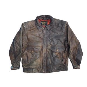 May include: A brown leather jacket with a collared neckline and two front pockets. The jacket has a ribbed hem and cuffs, and a red lining is visible at the collar. The jacket has a vintage, worn appearance.