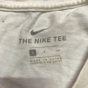 Op de afbeelding: Wit T-shirt met een Nike-logo en de tekst "THE NIKE TEE". Het label zegt "MADE IN CHINA FABRIQUE EN CHINE" en heeft een maat "S" label.