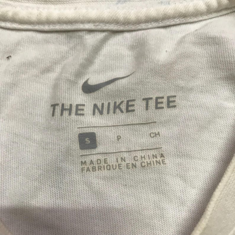 Op de afbeelding: Wit T-shirt met een Nike-logo en de tekst "THE NIKE TEE". Het label zegt "MADE IN CHINA FABRIQUE EN CHINE" en heeft een maat "S" label.
