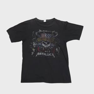 May include: Black t-shirt featuring a graphic design with a skull, an American flag, and the band name "Metallica." The design is distressed, giving it a vintage look. The shirt is short-sleeved and appears to be made of cotton.