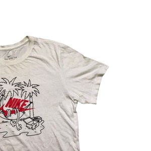 Op de afbeelding: Wit T-shirt met een zwart en rood Nike-logo en een afbeelding van een cartoonfiguur dat ontspant in een hangmat onder palmbomen. De tekst "THE NIKE TEE" is op het shirt gedrukt.