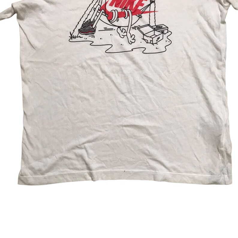 Op de afbeelding: Wit T-shirt met een cartoonillustratie van een Nike-schoen met een hoed en armen en benen. Het Nike swoosh-logo is rood en de tekst "NIKE" staat boven de schoen.