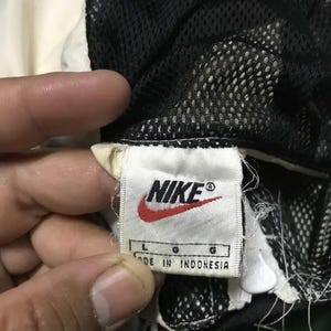 May include: Close-up of a Nike clothing tag. The tag is white with the Nike logo in red and black. The tag reads "L G MADE IN INDONESIA". The garment has a black mesh lining and a white and cream colored exterior.