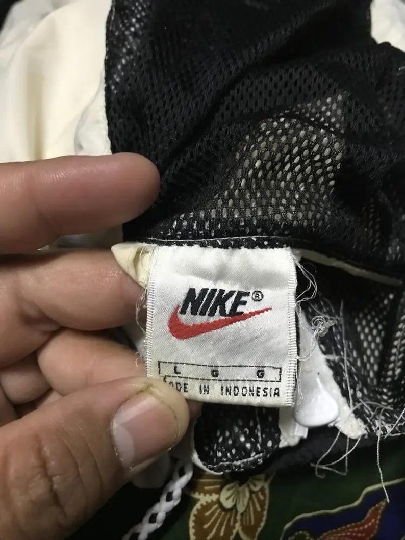 May include: Close-up of a Nike clothing tag. The tag is white with the Nike logo in red and black. The tag reads "L G MADE IN INDONESIA". The garment has a black mesh lining and a white and cream colored exterior.