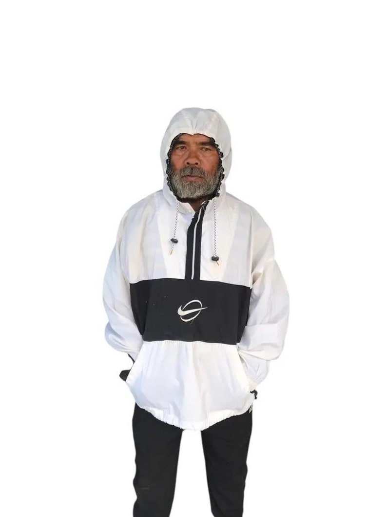 May include: White and black hooded anorak jacket with a front pocket and a black and white logo. The jacket has a drawstring hood and a half-zip closure. The model is wearing black pants.