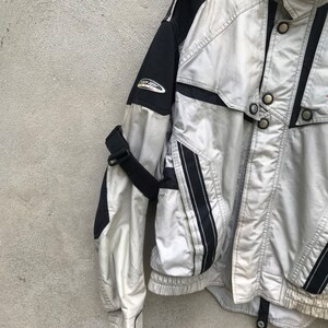K&ouml;nnte beinhalten: Eine silberne und schwarze Jacke mit Rei&szlig;verschluss und mehreren Druckkn&ouml;pfen. Die Jacke hat einen schwarzen Riemen am linken &Auml;rmel und einen schwarzen und silbernen Streifen an der rechten Seite.