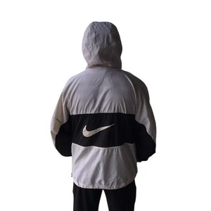 May include: A hooded jacket in white, black, and gray. The jacket features a large black panel across the back with a white Nike swoosh logo. The jacket has a full hood and is made of a lightweight material.