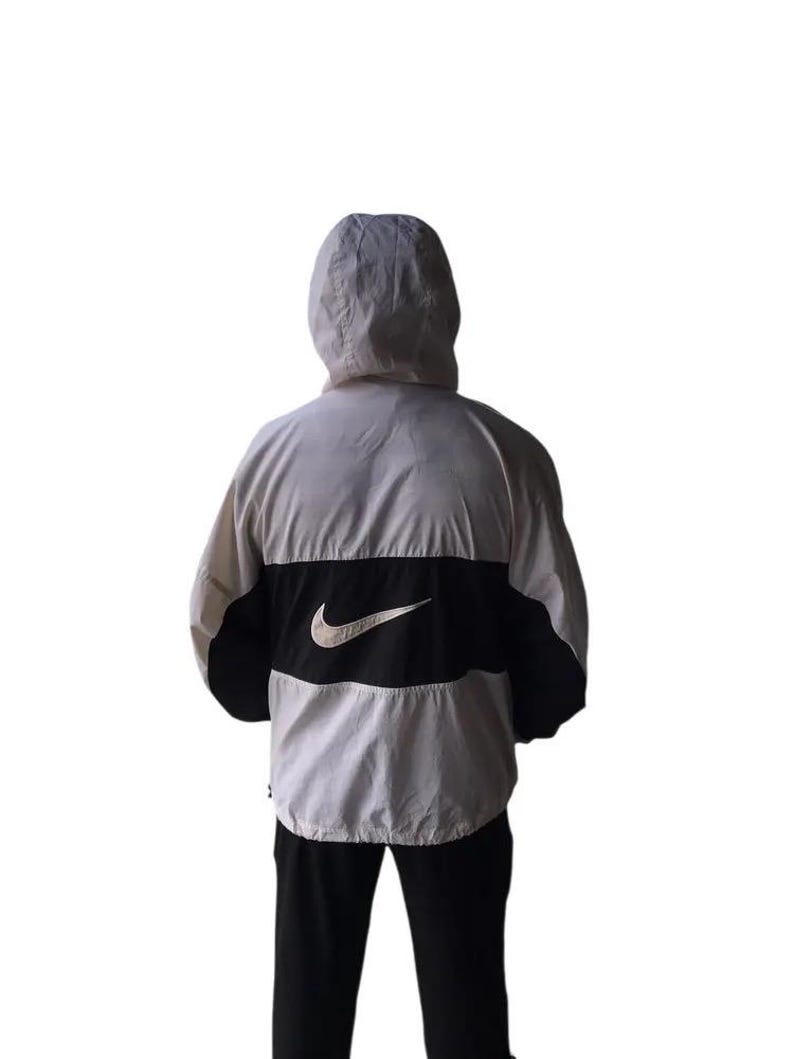 May include: A hooded jacket in white, black, and gray. The jacket features a large black panel across the back with a white Nike swoosh logo. The jacket has a full hood and is made of a lightweight material.