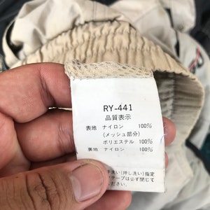 K&ouml;nnte beinhalten: Ein wei&szlig;es Etikett mit schwarzem Text, auf dem "RY-441" und "品質表示" (Qualit&auml;tsanzeige) steht. Das Etikett listet die Stoffzusammensetzung des Kleidungsst&uuml;cks auf: Nylon 100 % (Mesh-Teil), Polyester 100 %, Nylon 100 %. Das Etikett enth&auml;lt auch Waschanweisungen: "Handw&auml;sche (Pressw&auml;sche) vorgeschrieben. Klebeband muss geschlossen sein."