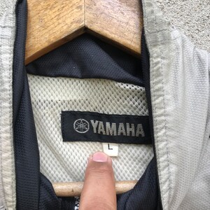 K&ouml;nnte beinhalten: Eine graue Yamaha-Jacke mit schwarzem Mesh-Futter und einem schwarzen Etikett mit dem Yamaha-Logo und dem Buchstaben "L" darauf.