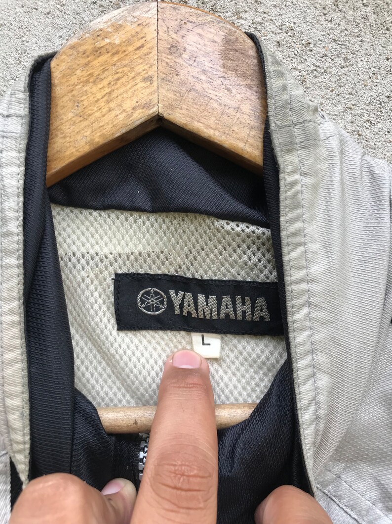 K&ouml;nnte beinhalten: Eine graue Yamaha-Jacke mit schwarzem Mesh-Futter und einem schwarzen Etikett mit dem Yamaha-Logo und dem Buchstaben "L" darauf.