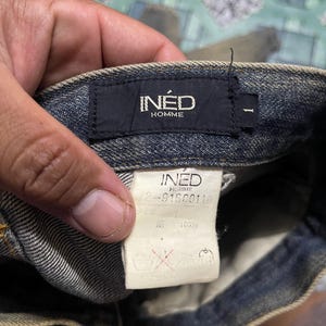 May include: Close-up of a pair of blue denim jeans. The black label reads "IN&Eacute;D HOMME". A white tag displays the brand name and product information. The jeans are made of 100% cotton.