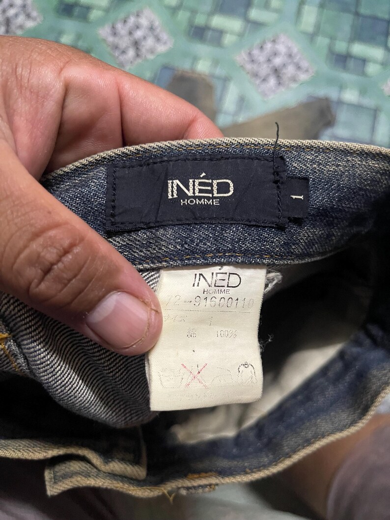 May include: Close-up of a pair of blue denim jeans. The black label reads "IN&Eacute;D HOMME". A white tag displays the brand name and product information. The jeans are made of 100% cotton.