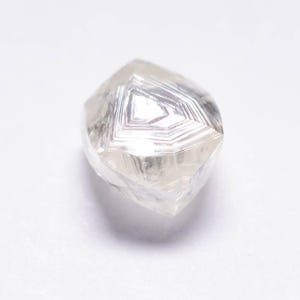Op de afbeelding: Een heldere, ongeslepen diamant met een geometrisch patroon van facetten. De diamant heeft een lichtgrijze kleur en een kristallijne structuur. De edelsteen is tegen een witte achtergrond geplaatst.