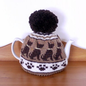 Könnte beinhalten: Ein brauner und weißer gestrickter Teekannenwärmer mit einem braunen Bommel oben. Der Teekannenwärmer hat ein Muster aus Pfotenabdrücken und schwarzen Katzen.